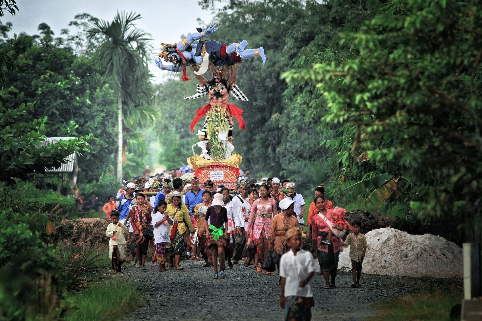 An ogoh-ogoh borne along a village path through the trees, the procession stretching behind it.