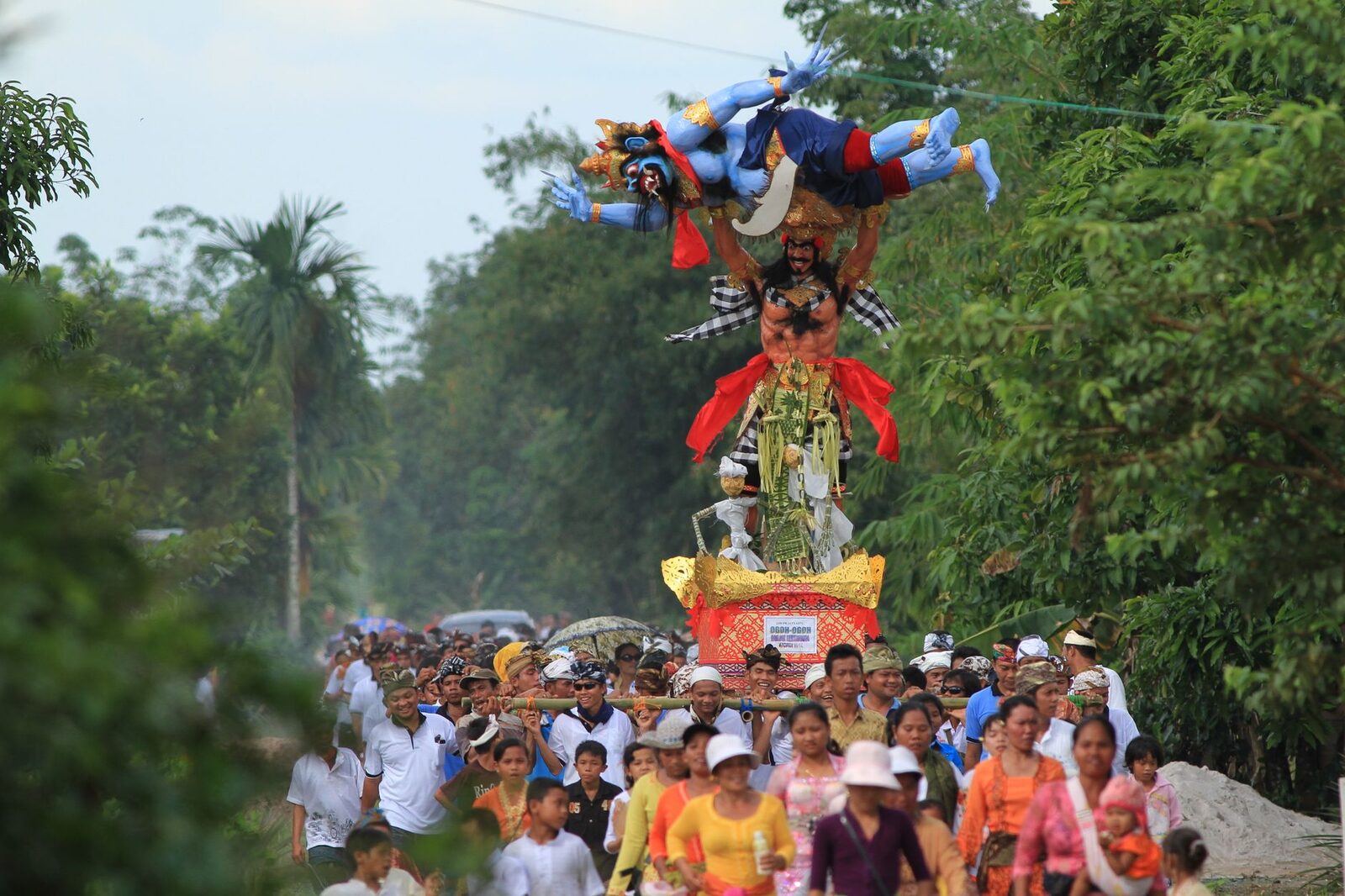 A colourful ogoh-ogoh borne along the road on its bamboo frame by a village crowd.