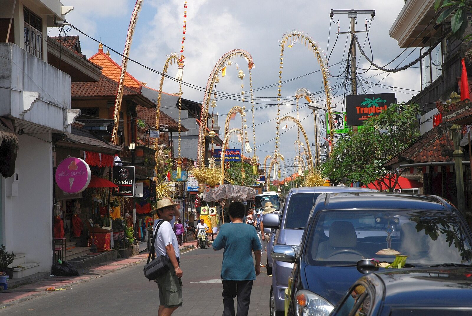 Penjor arching over a street for Galungan — tall bamboo poles hung with coconut leaf, fruit and rice, going up all along the road.