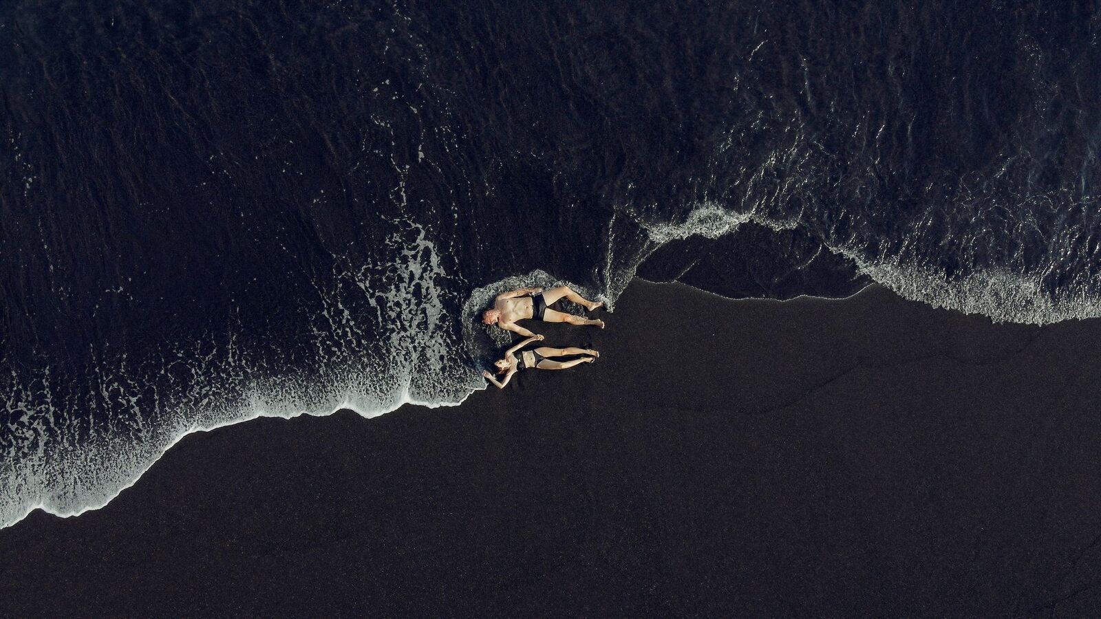 From above, dark sand meeting the sea. (A Bali black-sand beach.)