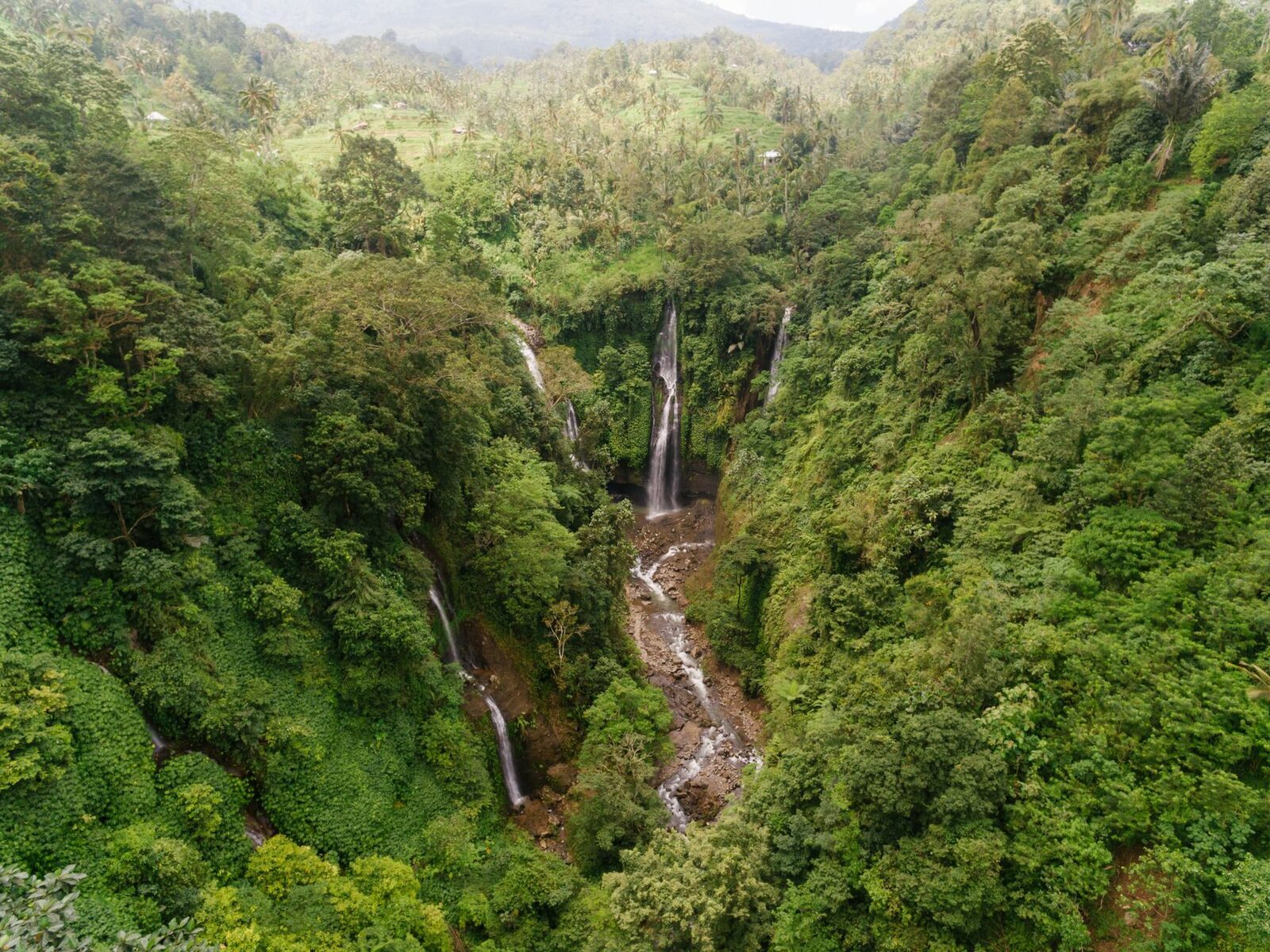 Aerial over a cascade threading a green valley — the country the waterfall loops string together. (A north-Bali jungle waterfall.)