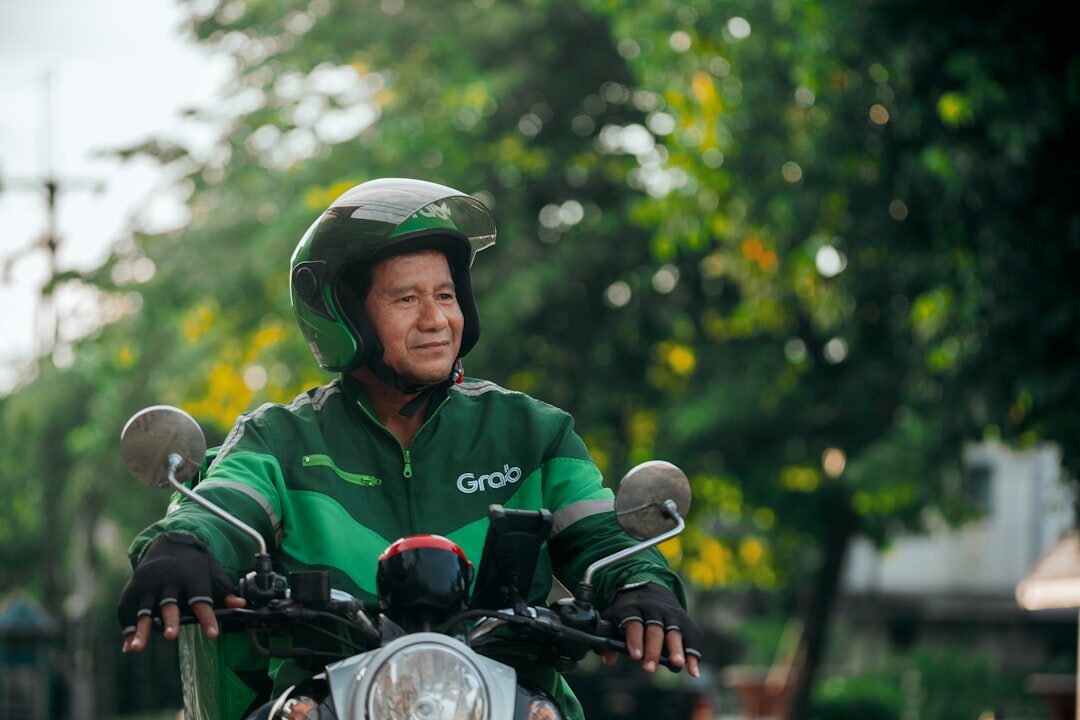 A Grab driver in the green jacket and helmet waits on his scooter — ride-hailing is the no-stress alternative to riding yourself.