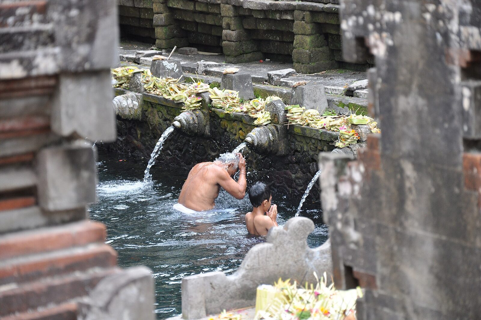 A worshipper drinking and washing at a single spout, offerings floating past.